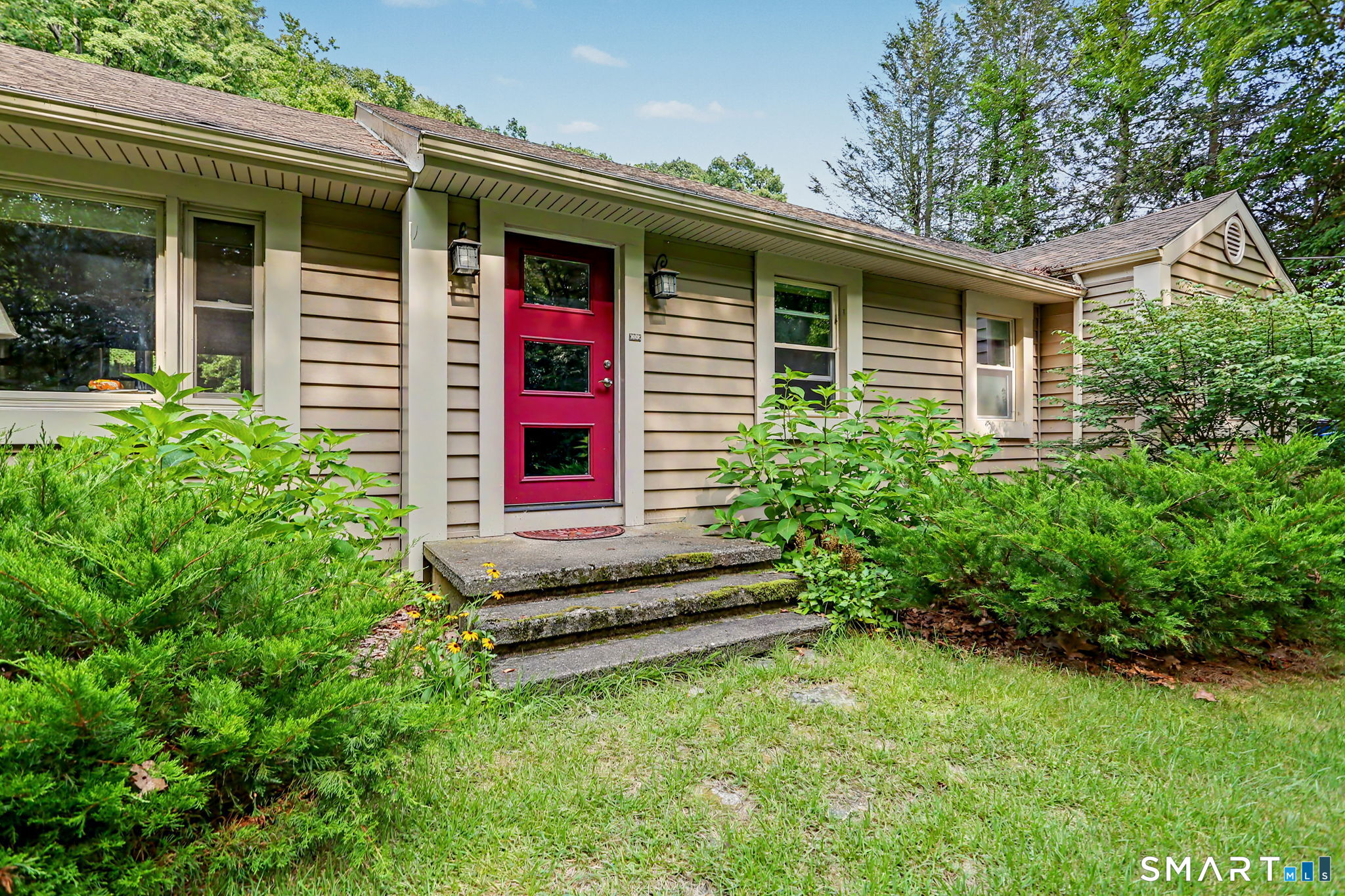 Single-story beige home with a bright red front door, concrete steps, and lush shrubs.