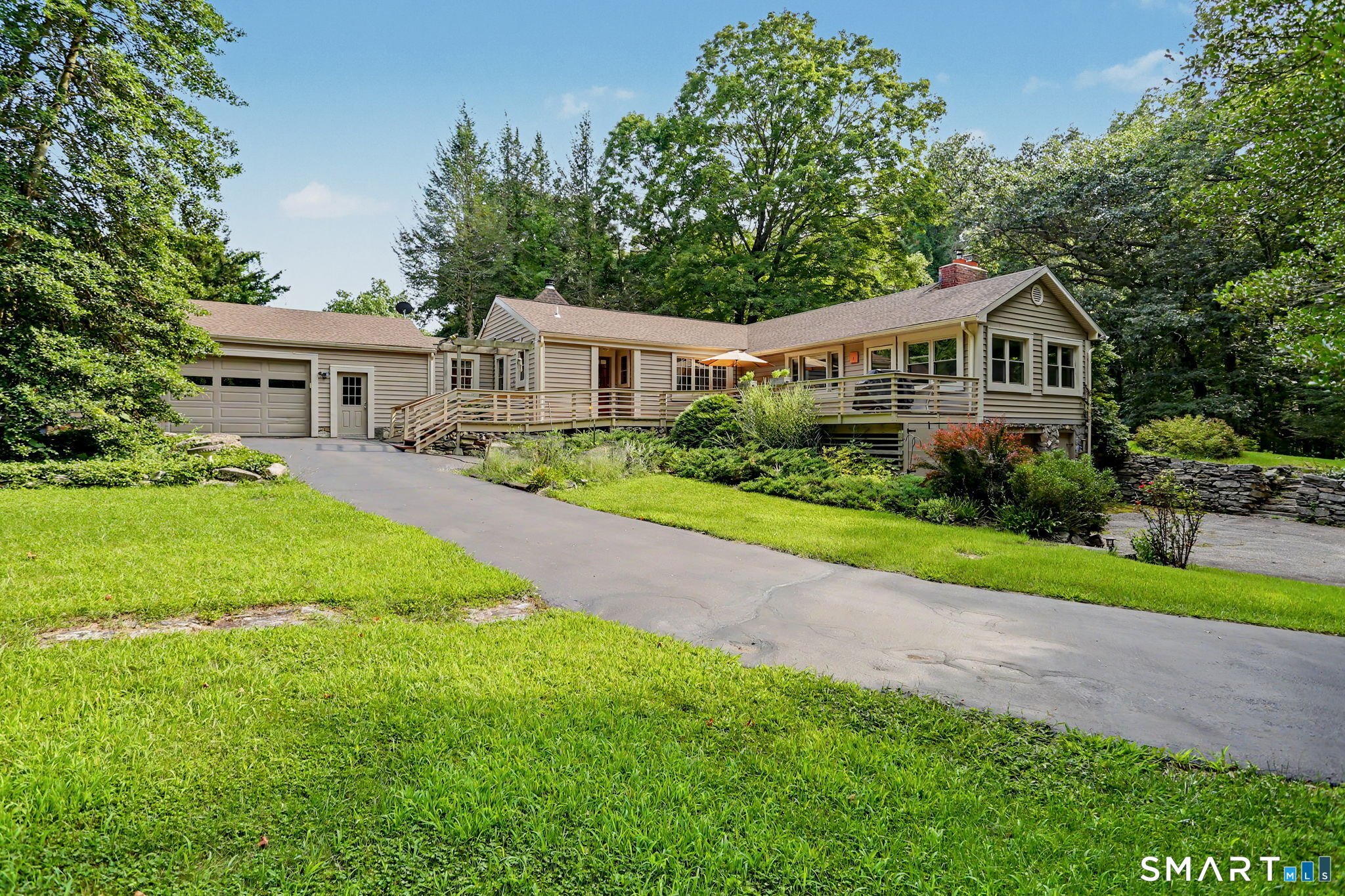Single-story beige home with an attached garage, raised deck, and tree-lined lawn.