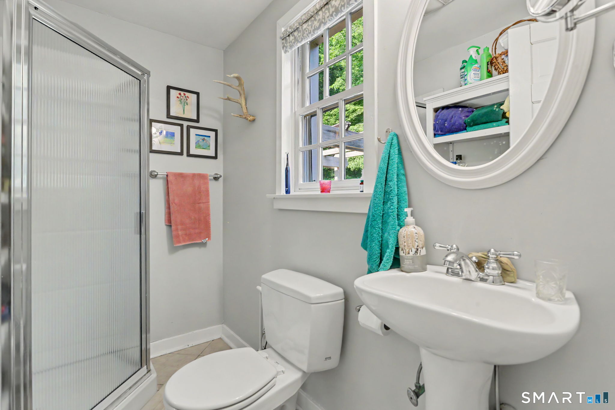 Bathroom with a glass shower, pedestal sink, oval mirror, and window overlooking green trees.