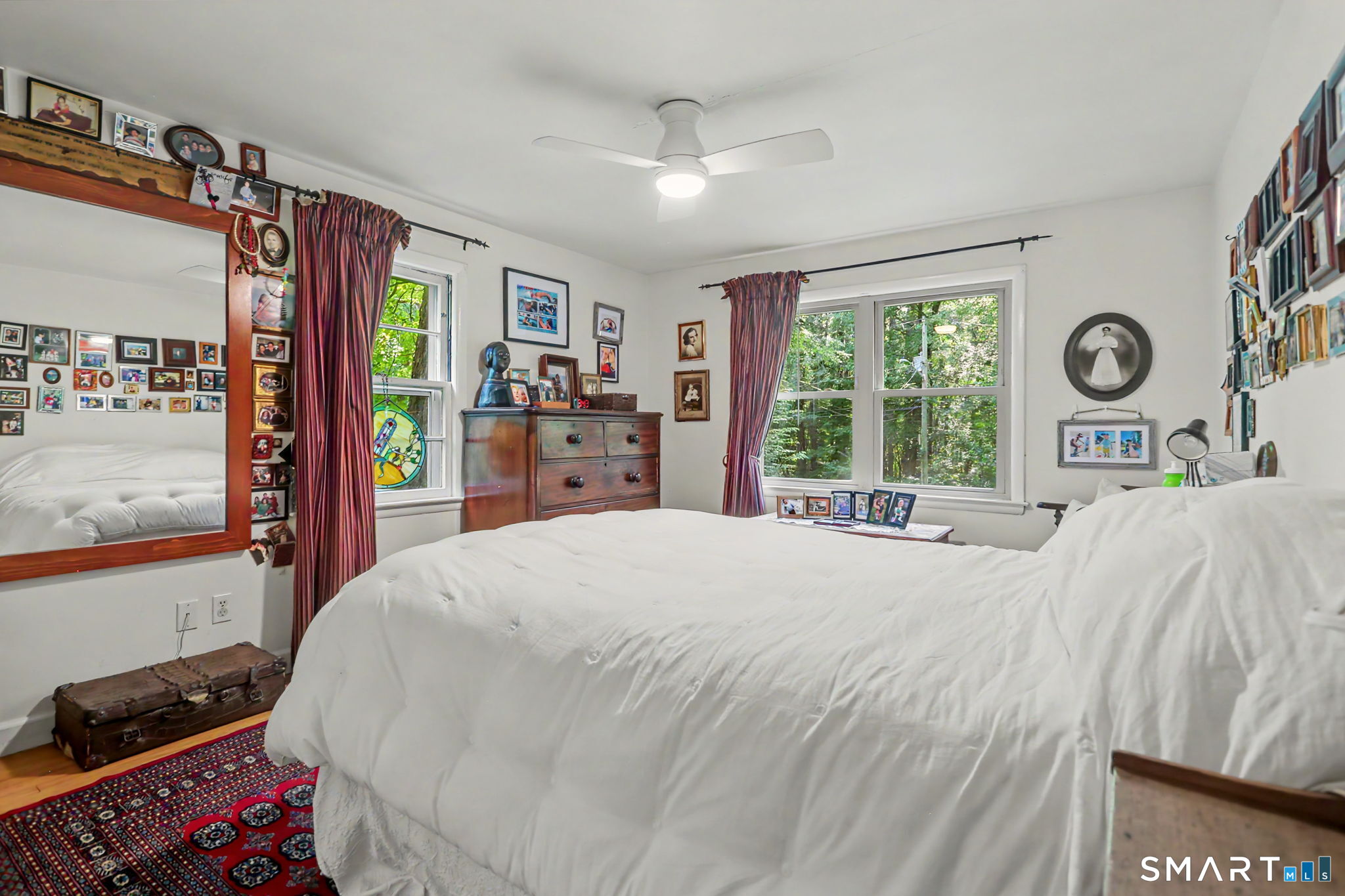 Bedroom with a large white bed, burgundy curtains, a wooden dresser, and leafy views through the windows.