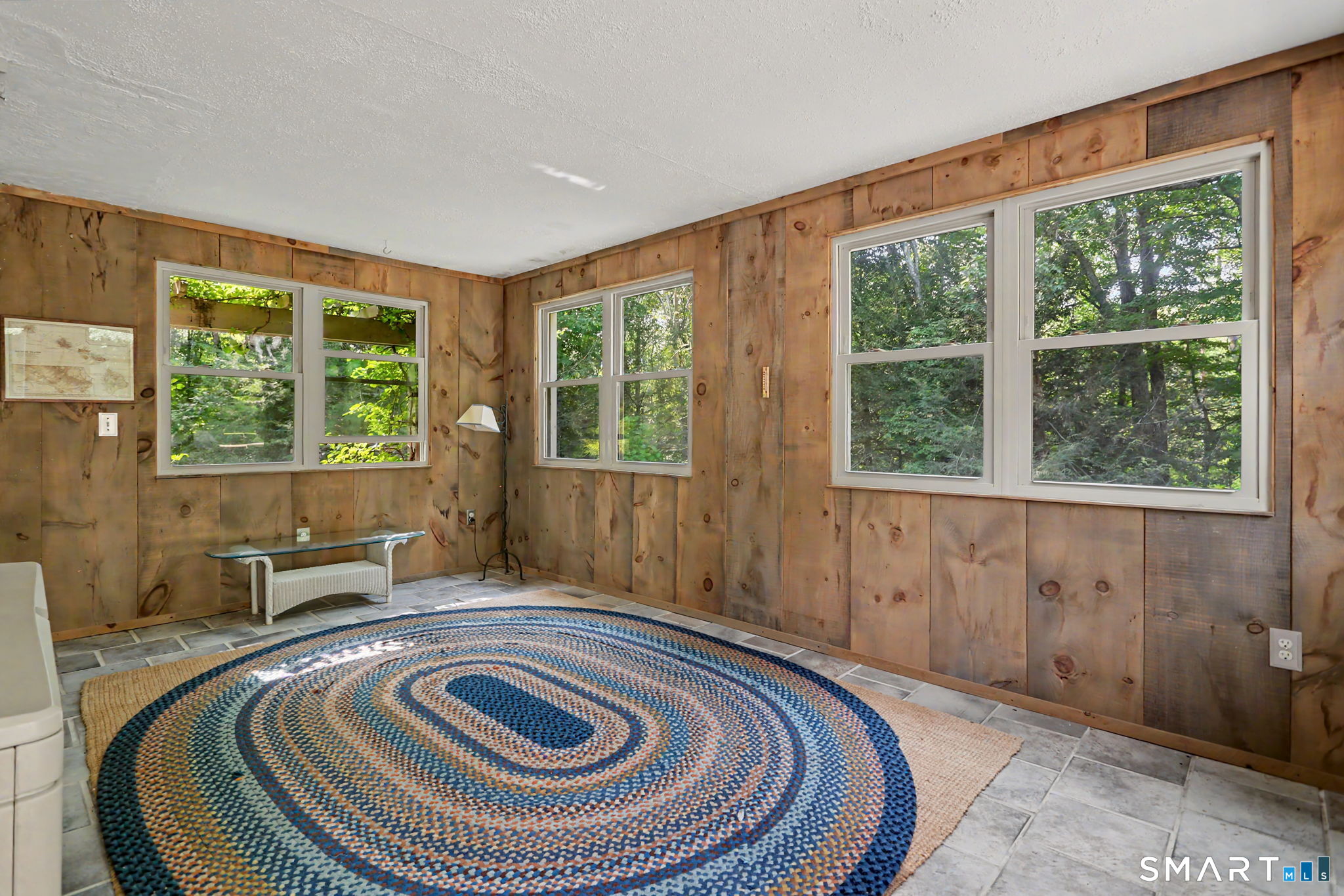 Wood-paneled room with large windows overlooking trees and a blue braided rug.
