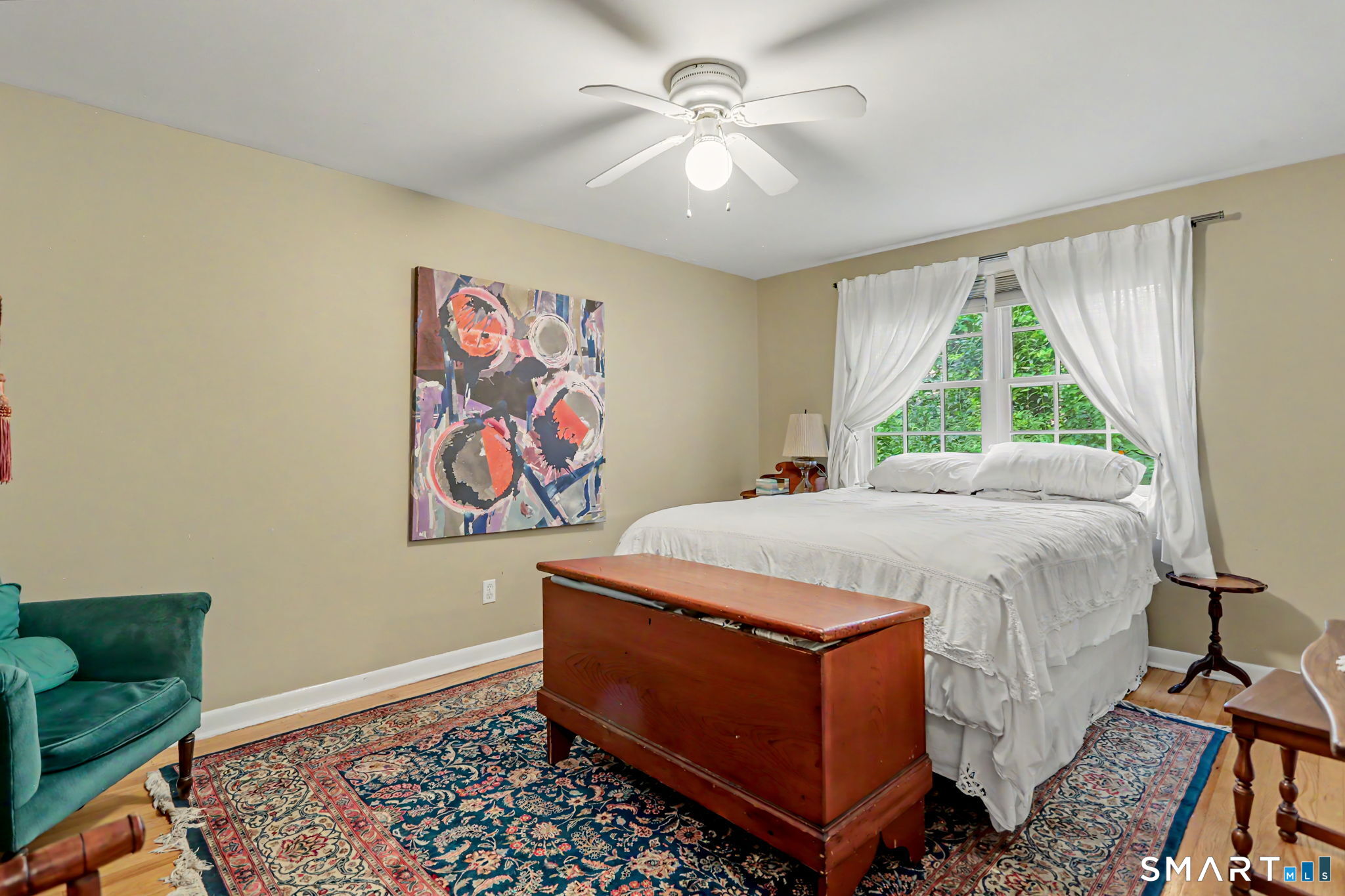 Bedroom with a white bed, curtained windows, wooden blanket chest, and patterned rug.