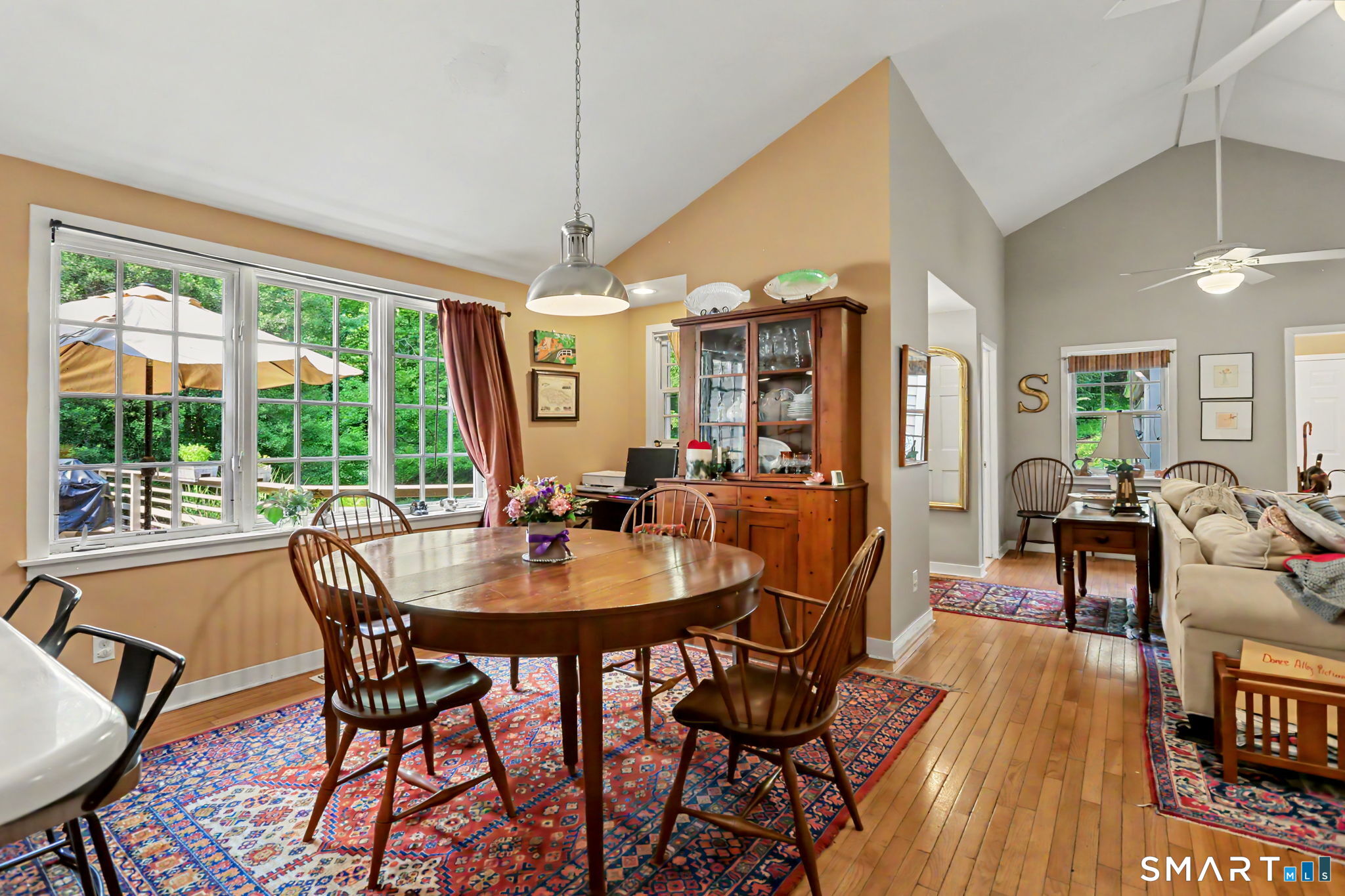 Dining area with a wooden table and large windows opening onto greenery, beside a furnished living room.