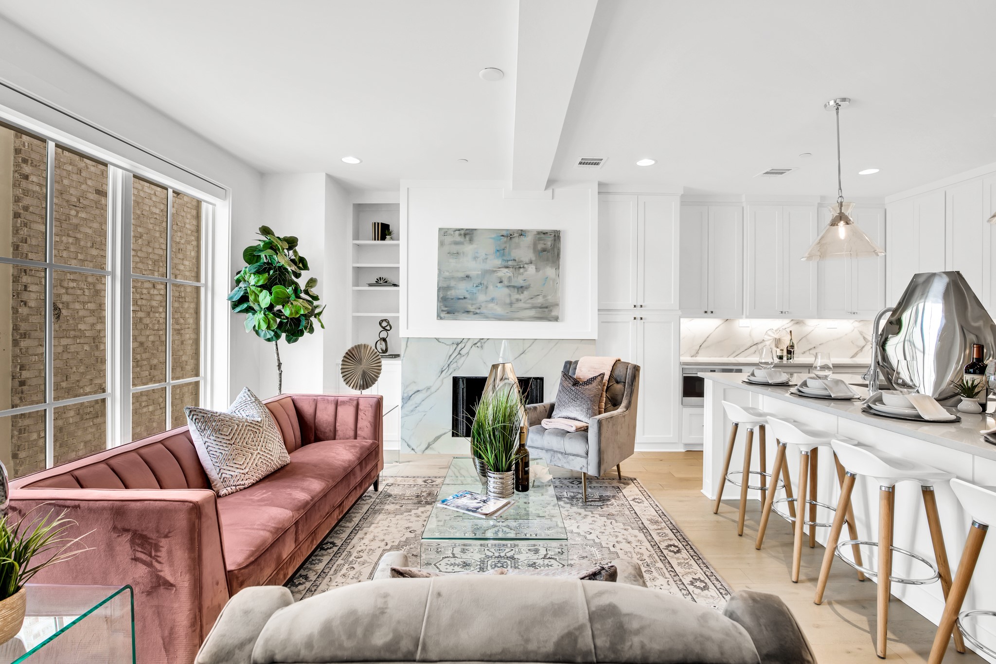 Open-plan living room and kitchen with a pink sofa, marble fireplace, and white island seating.