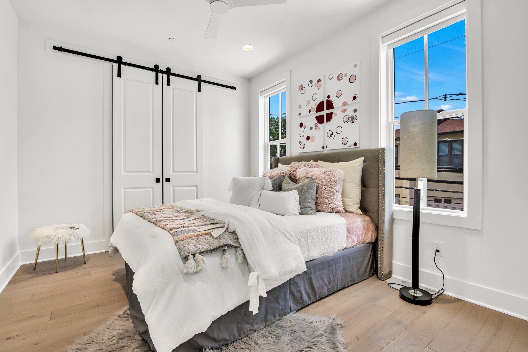 Staged bedroom with white bedding, a gray upholstered headboard, sliding barn doors, and tall windows.