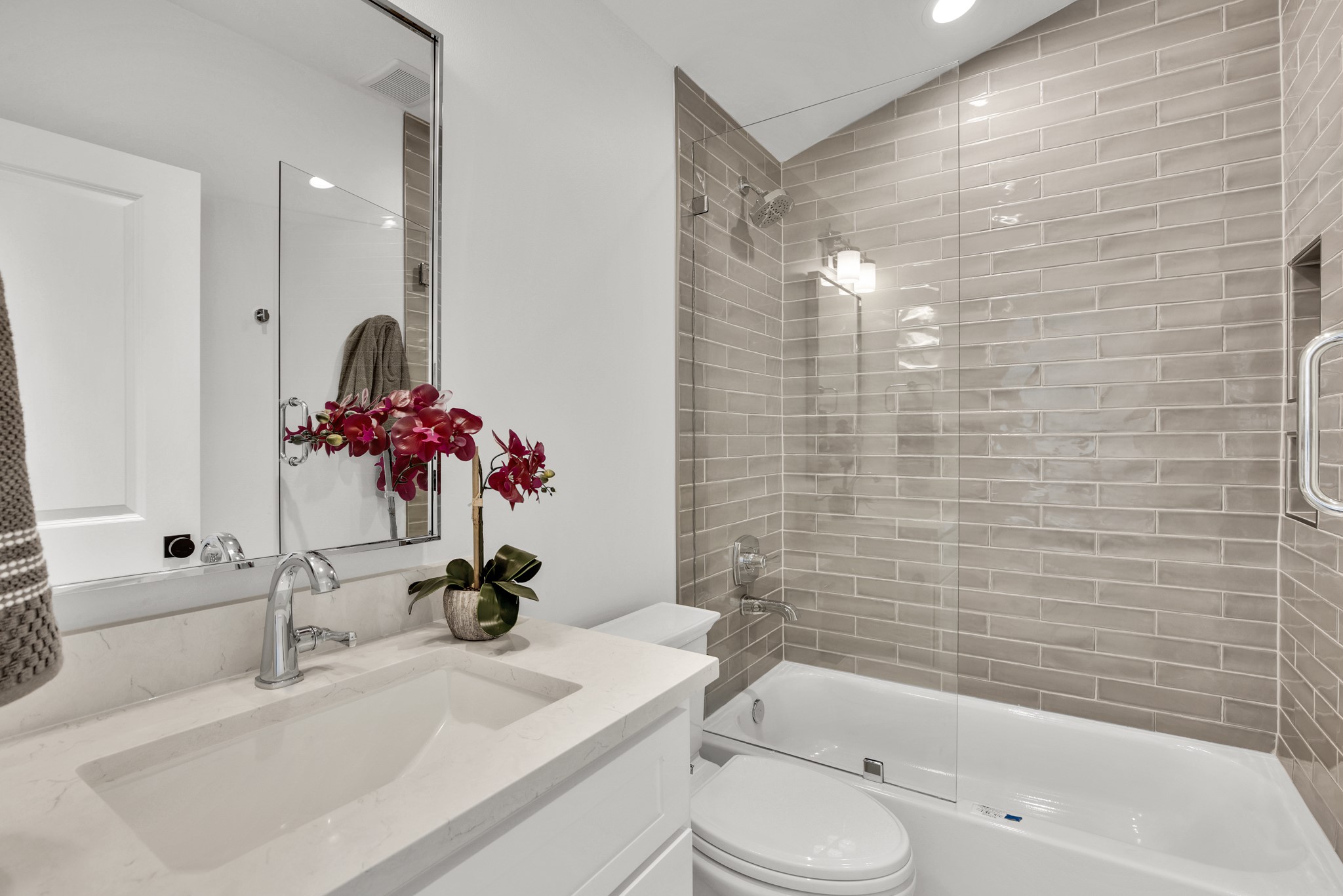 Bathroom with gray tiled tub and glass shower beside a marble vanity topped with a red orchid.