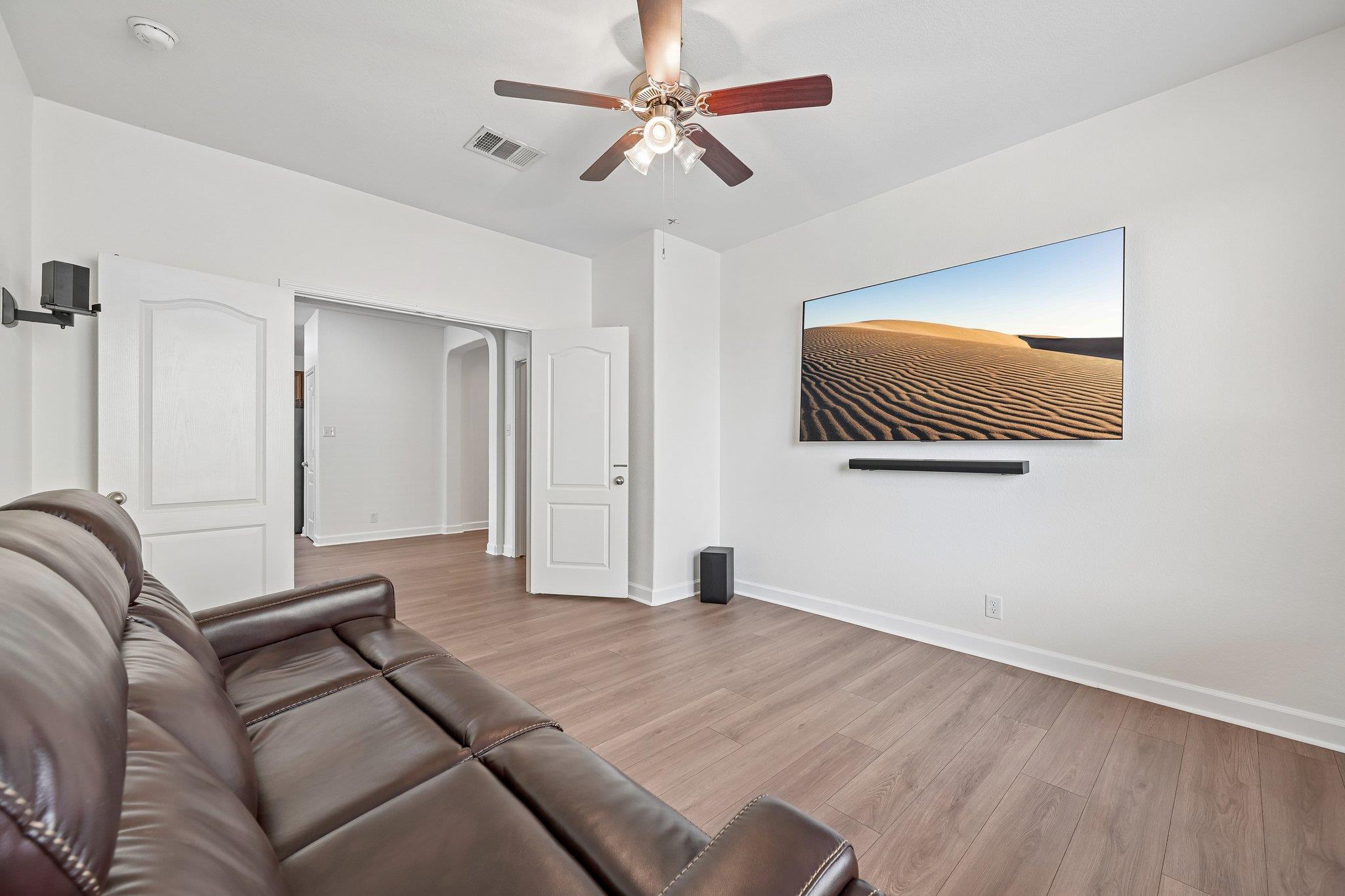 Living room with a leather sectional, wood-look floors, ceiling fan, and wall-mounted television.