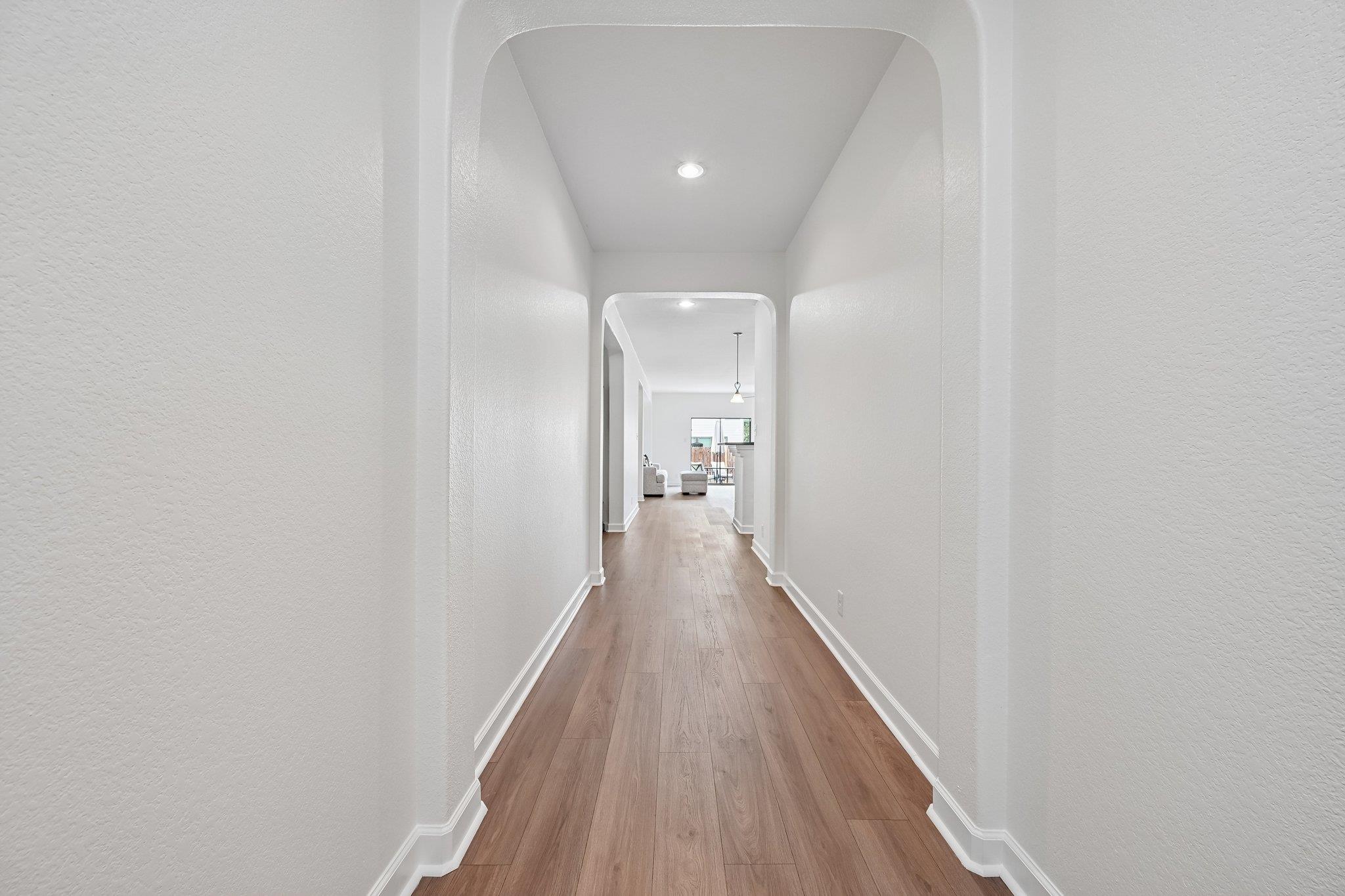 Bright hallway with arched openings, recessed lights, and warm wood flooring leading to a living area.