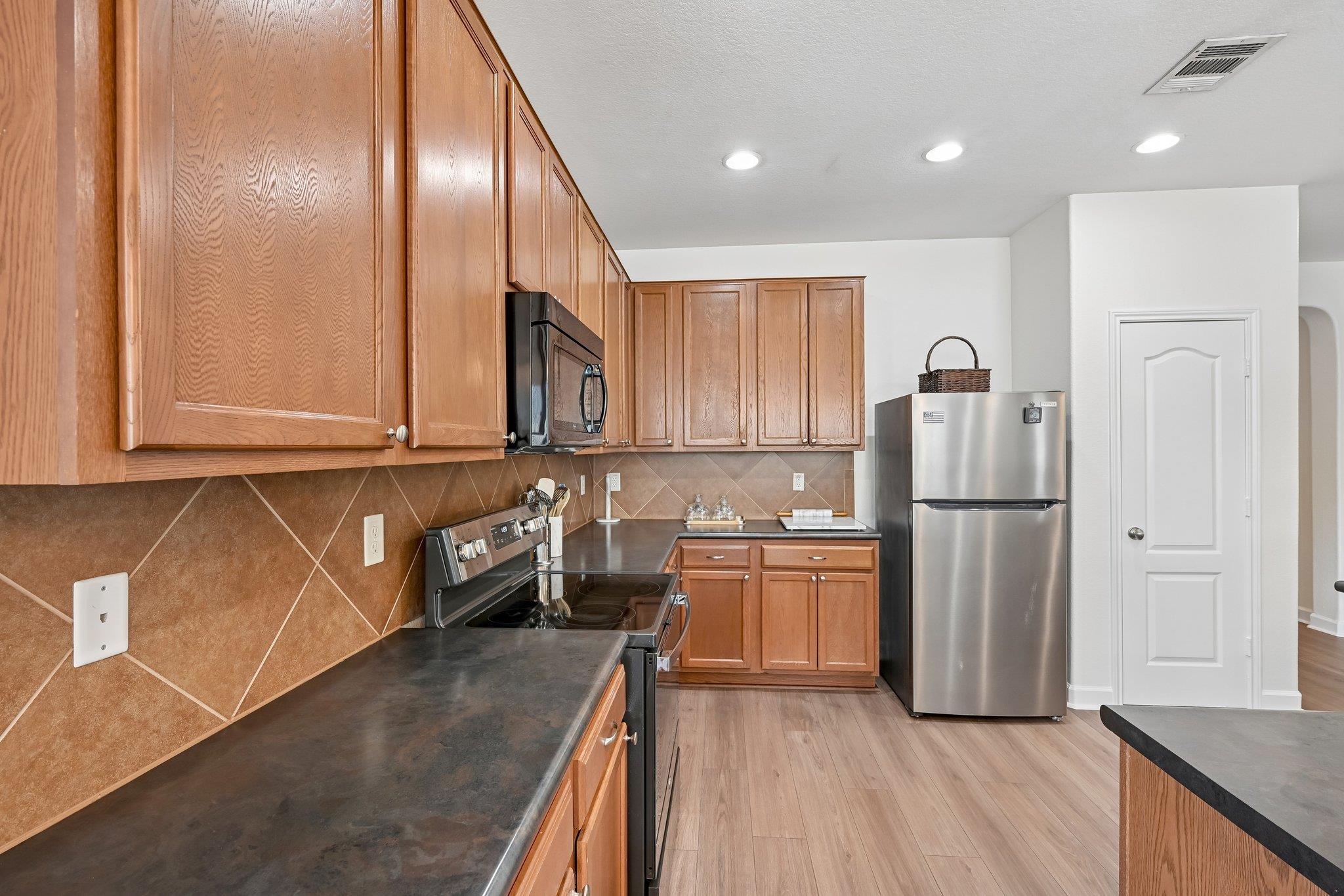 Kitchen with wood cabinets, brown tile backsplash, dark counters, and stainless steel refrigerator.