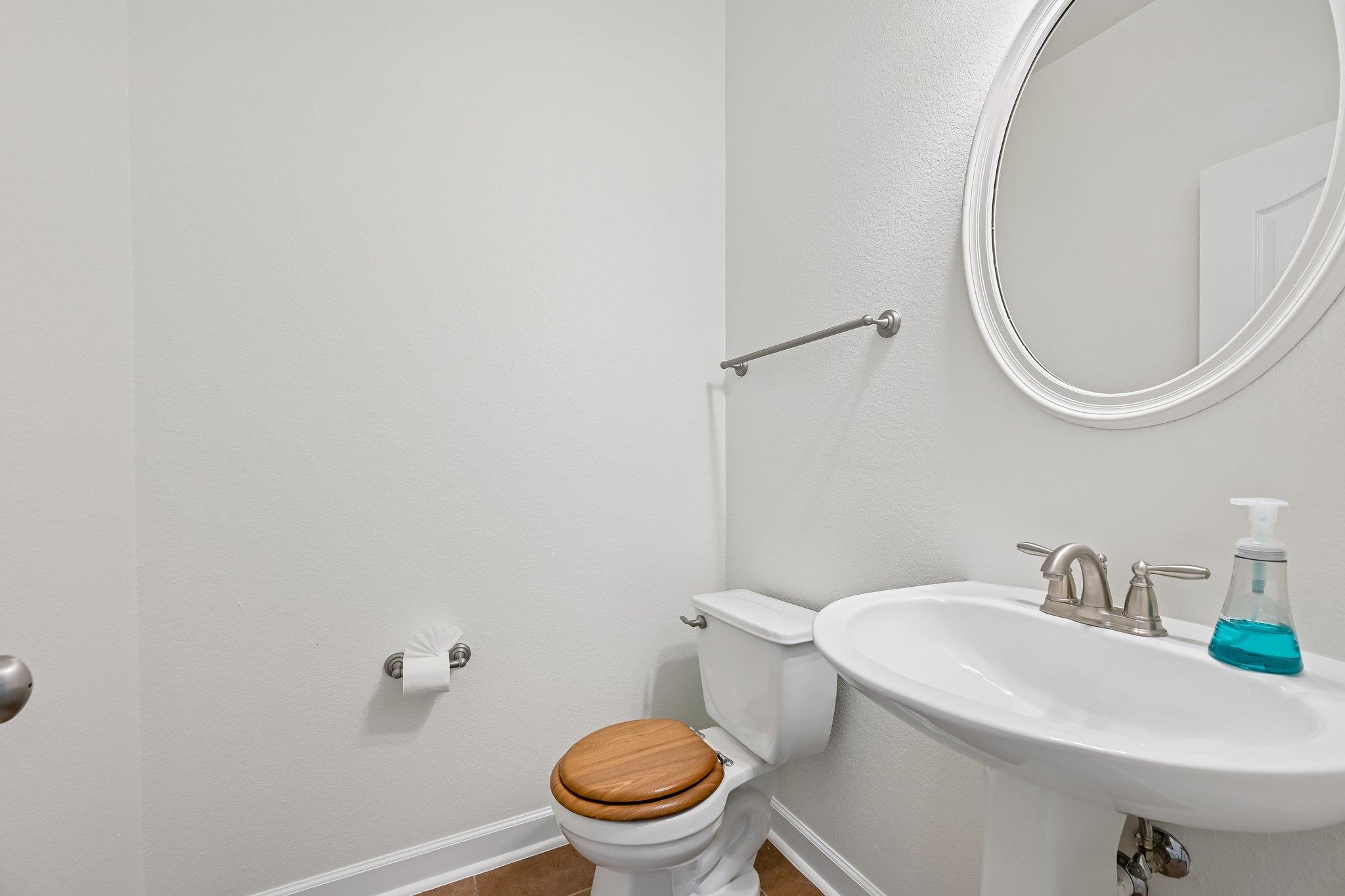 Bathroom with a wood-seat toilet, pedestal sink, and large round mirror.