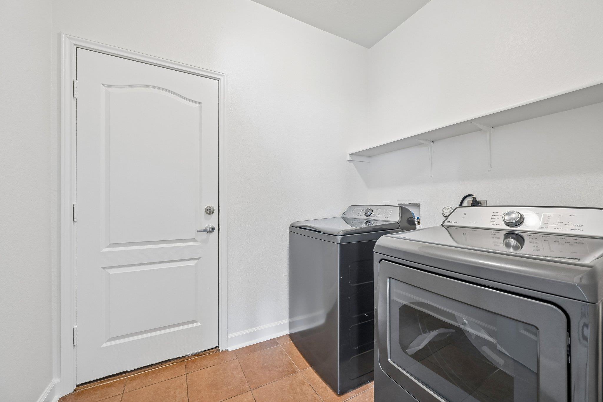 Laundry room with gray washer and dryer, wall shelf, and terracotta tile floor.