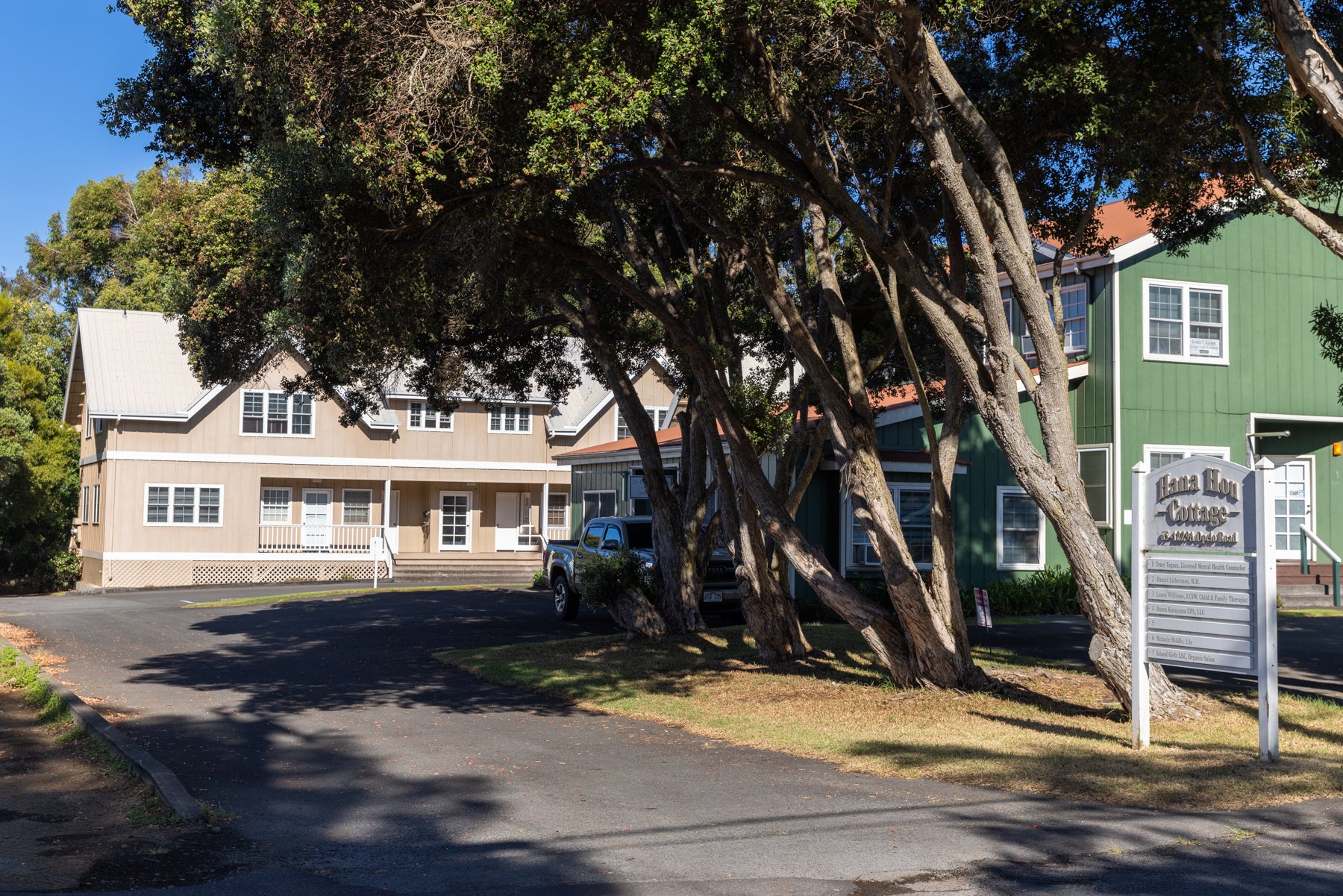 Tan and green buildings beneath large shade trees, with a Hana Hou Cottage directory sign.