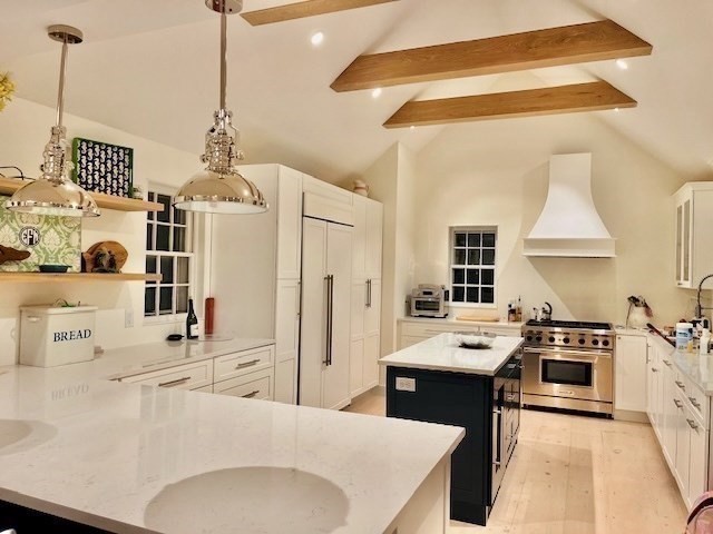 White kitchen with a dark island, stainless steel range, pendant lights, and exposed wooden ceiling beams.