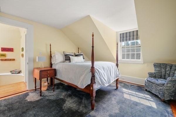 Bedroom with a four-poster bed, sloped ceiling, striped window valance, and patterned armchair.