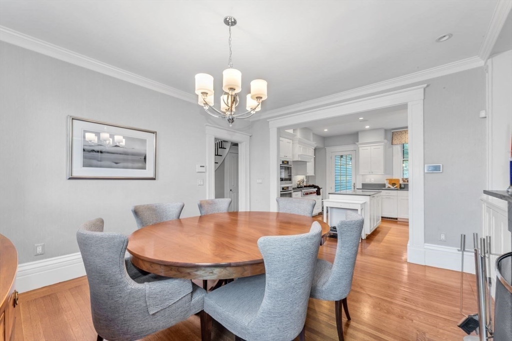 Dining room with a round wooden table, gray chairs, and a chandelier beside an open kitchen.