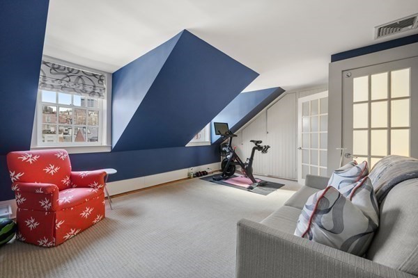 Attic sitting room with a blue angled wall, red floral chair, gray sofa, and exercise bike.
