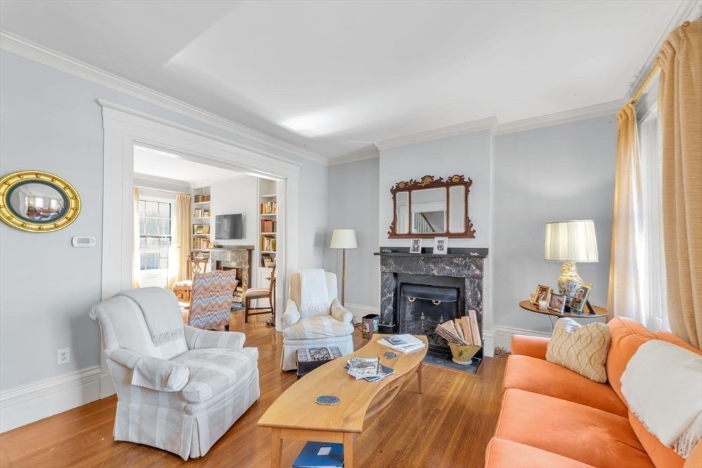 Living room with a marble fireplace, hardwood floors, white chairs, and an orange sofa.