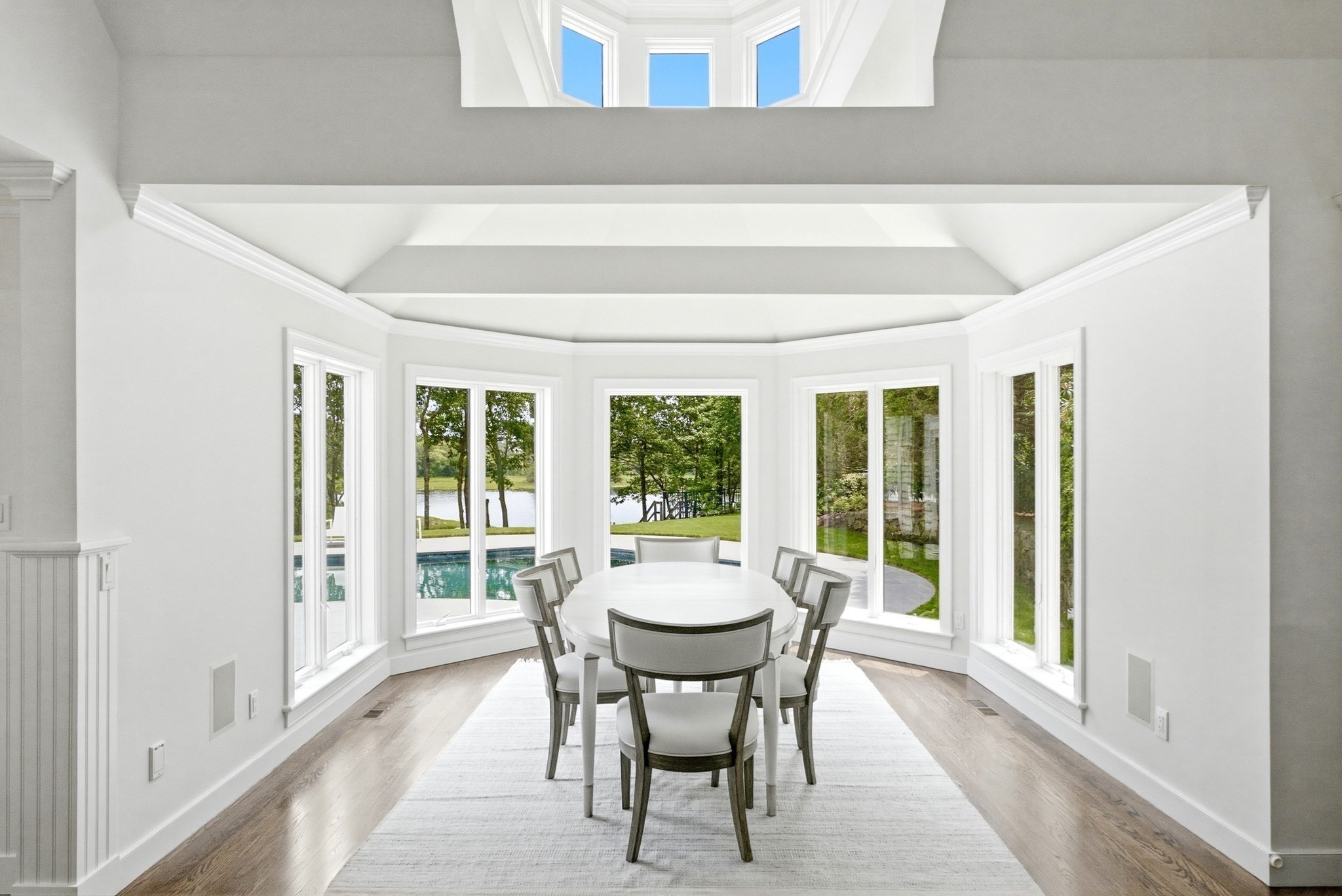 Dining room with a vaulted ceiling, oval table, and windows overlooking a pool and water.