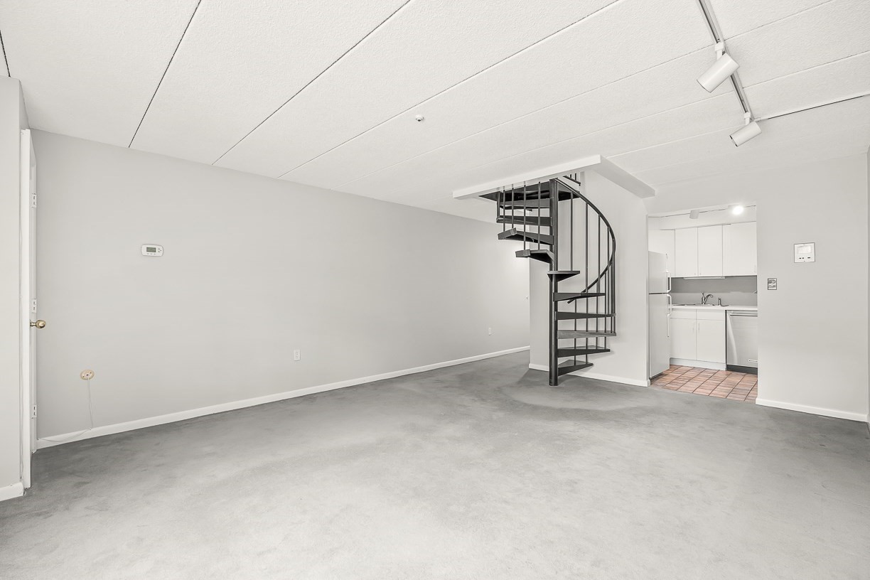 Empty condo living area with a black spiral staircase and compact white kitchen.