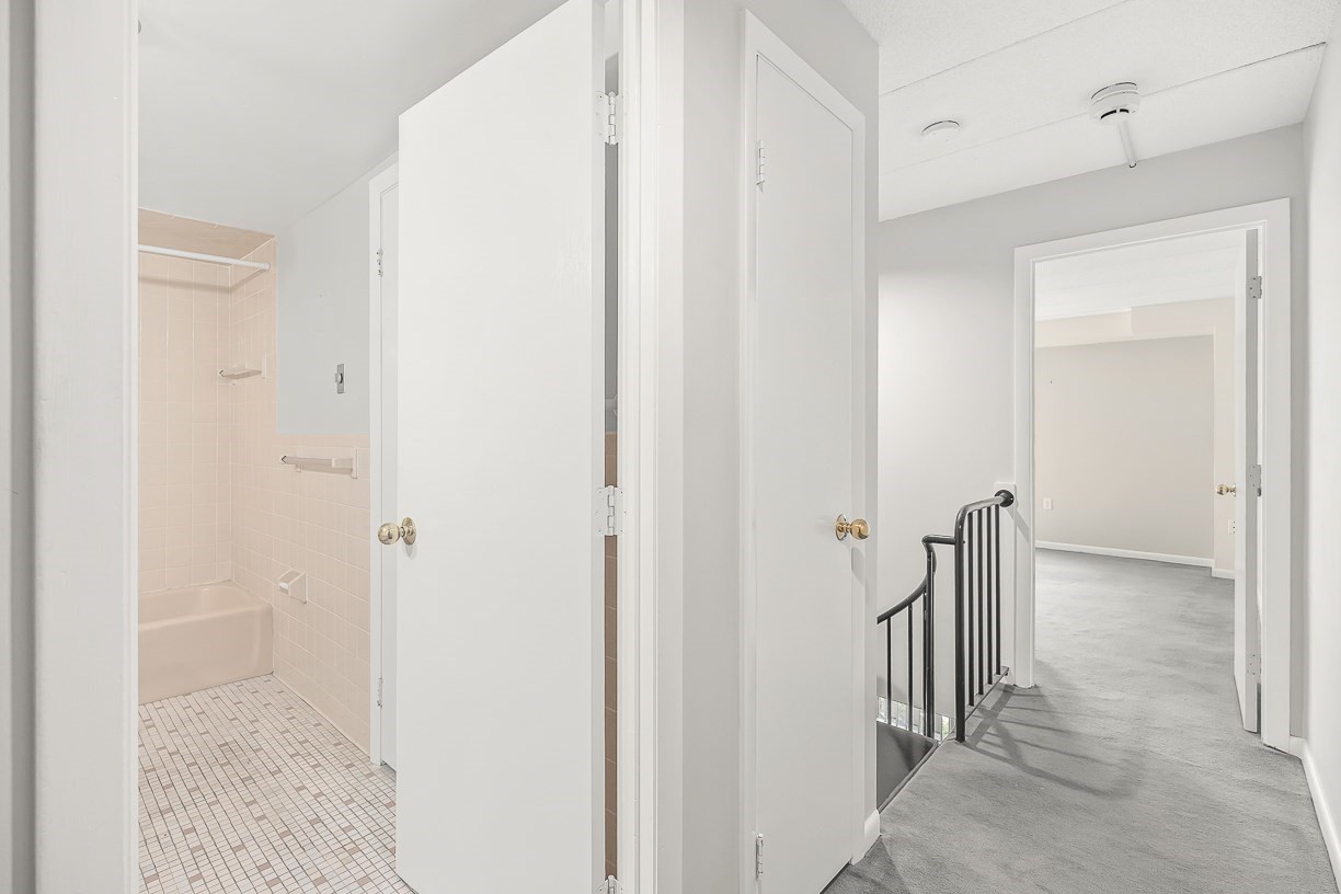 Apartment hallway with gray carpet, black stair railing, and a pink-tiled bathroom.