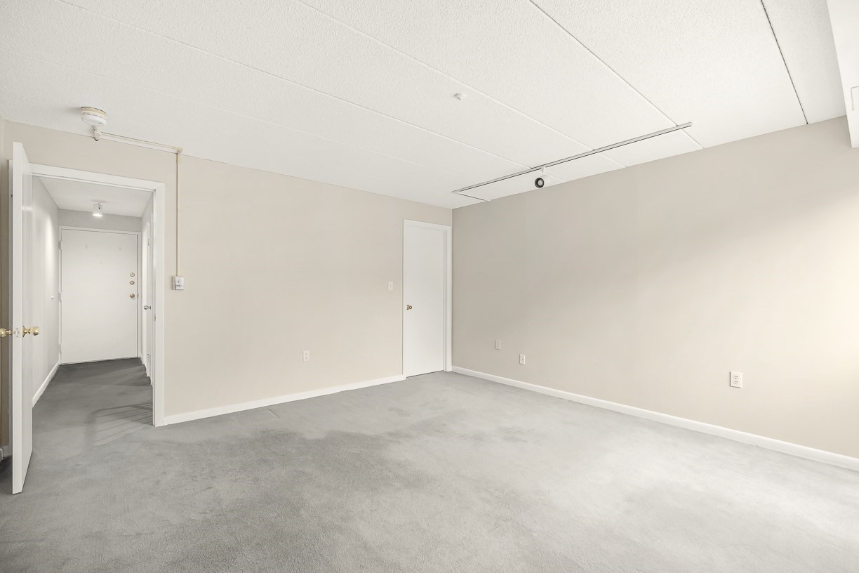 Empty condo room with gray carpeting and doors to the hallway and adjoining room.