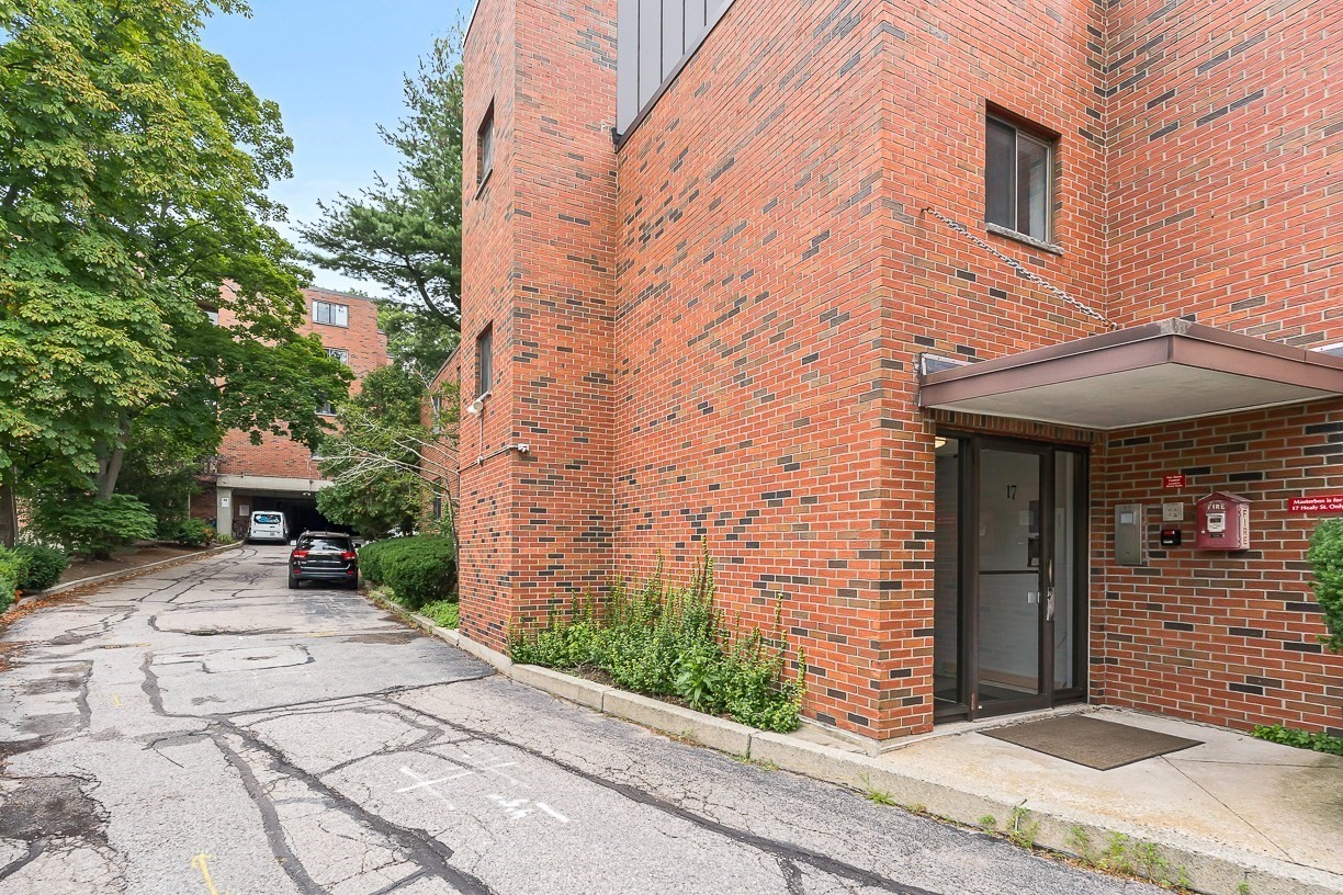 Brick apartment building entrance with glass door marked 17, metal canopy, and driveway beside leafy trees.
