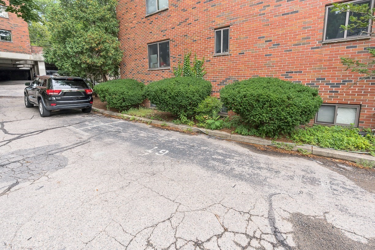 Red-brick apartment building beside a cracked parking area with shrubs and a parked SUV.