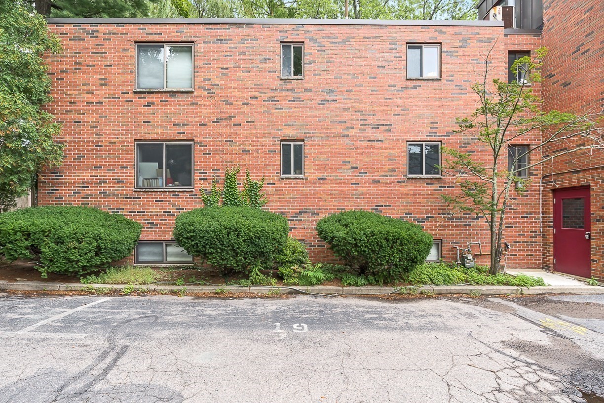 Red brick apartment building with trimmed shrubs, a red entrance door, and parking area.