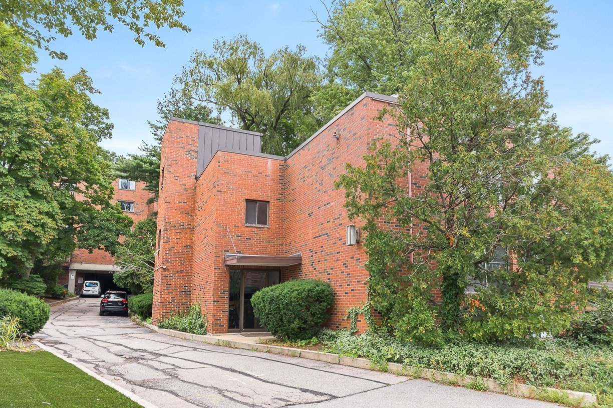 Red-brick condominium building with a recessed entrance, mature trees, and a driveway.