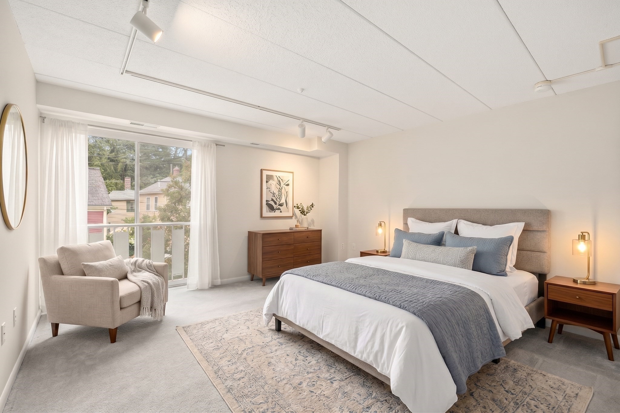 Staged bedroom with a gray upholstered bed, wood nightstands, and an armchair beside a glass door.