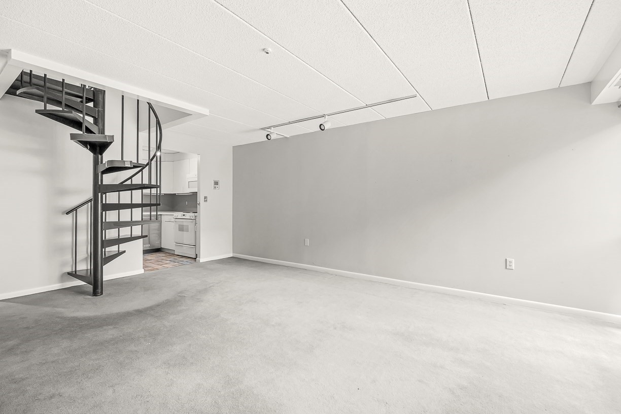 Vacant living area with gray carpet, a black spiral staircase, and a compact kitchen.