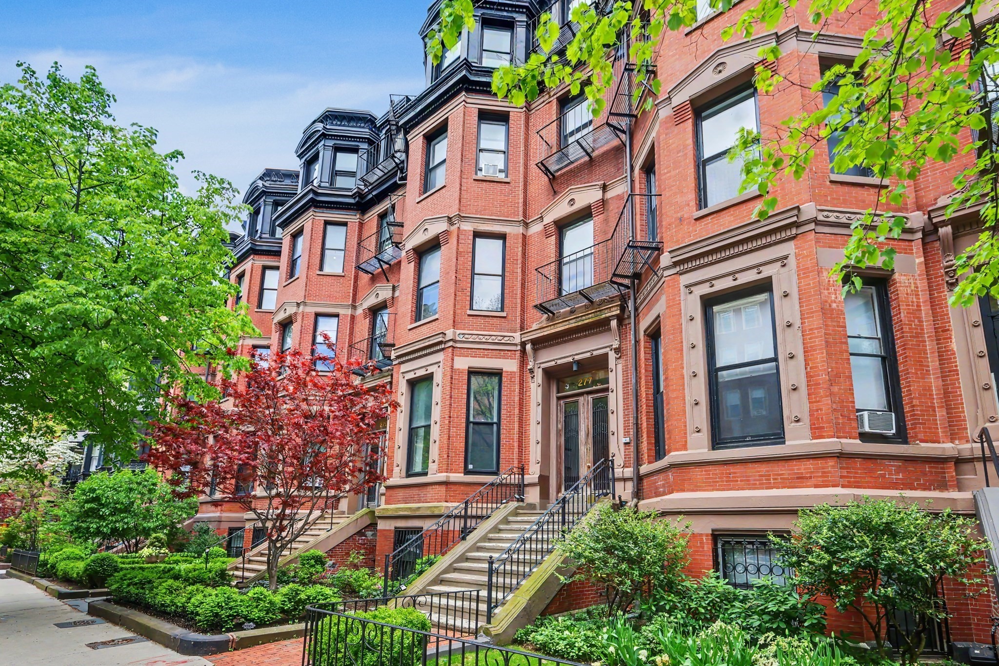 Red-brick Boston townhouse with ornate trim, black fire escapes, front stoops, and leafy landscaping.
