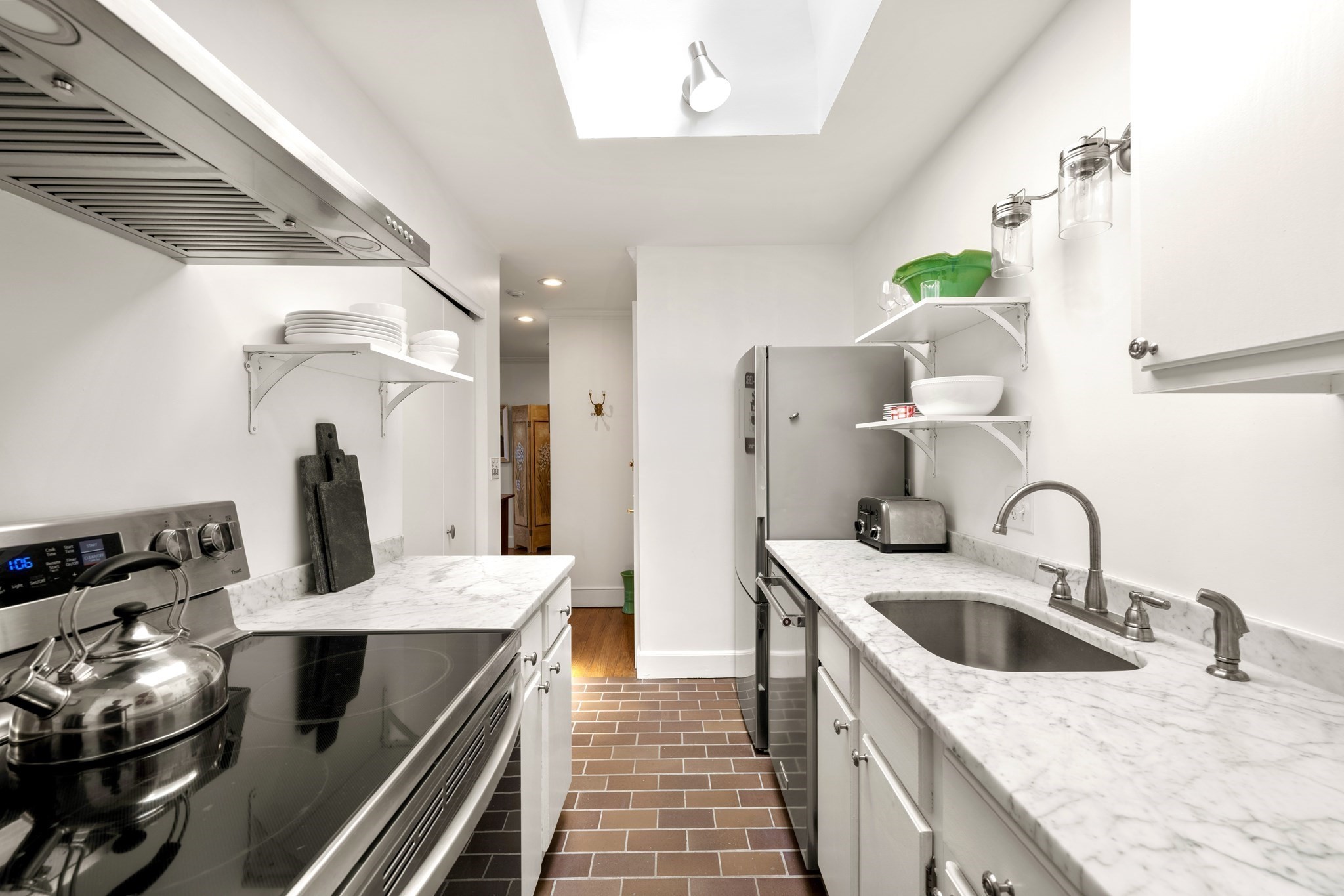Narrow white galley kitchen with marble counters, stainless steel appliances, and brick tile flooring.