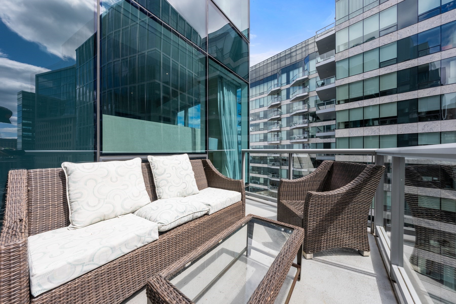 Furnished balcony with wicker seating and glass railing overlooking modern high-rise buildings.