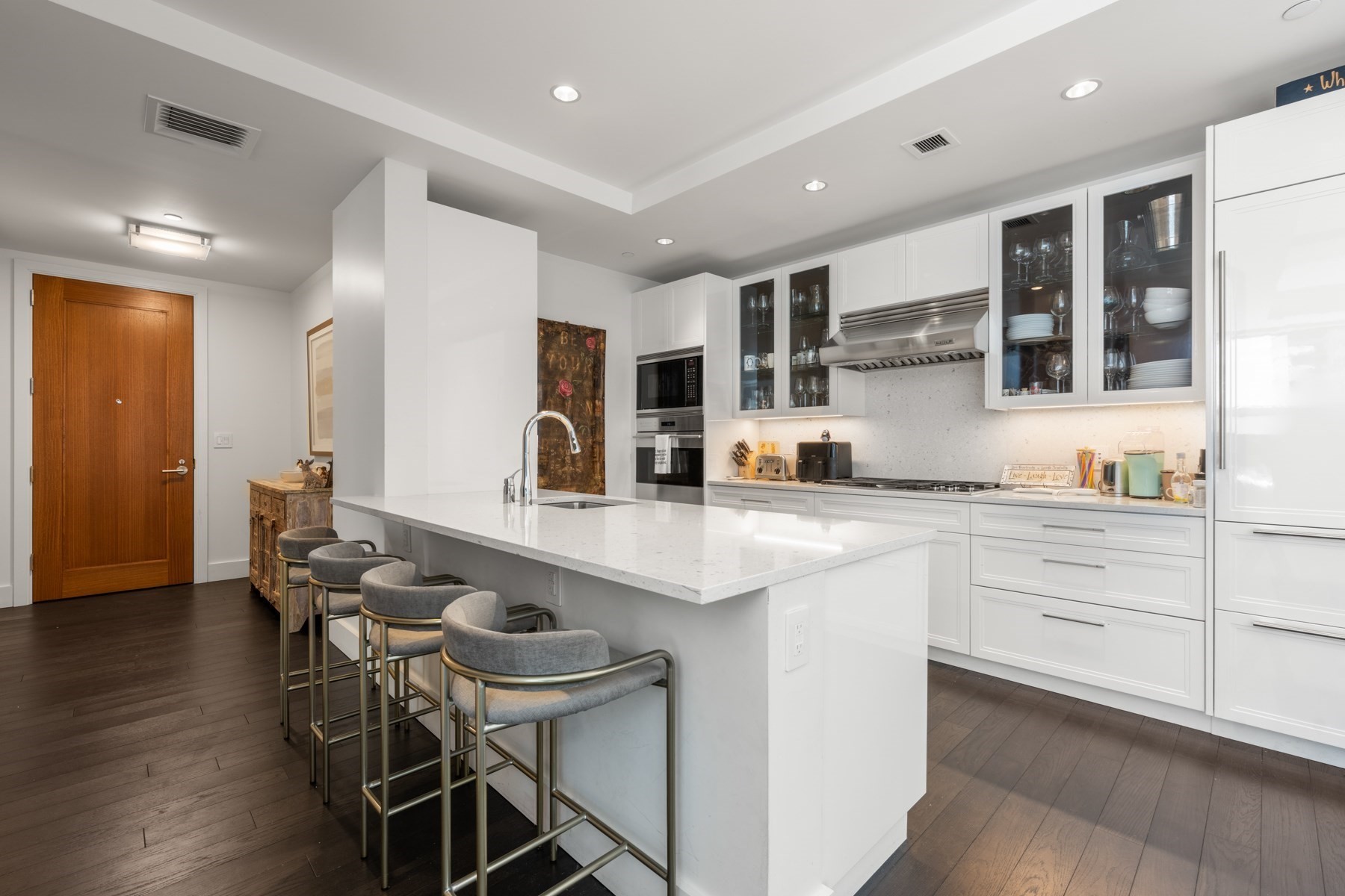 Modern white kitchen with a large island, four gray stools, and glass-front cabinets.