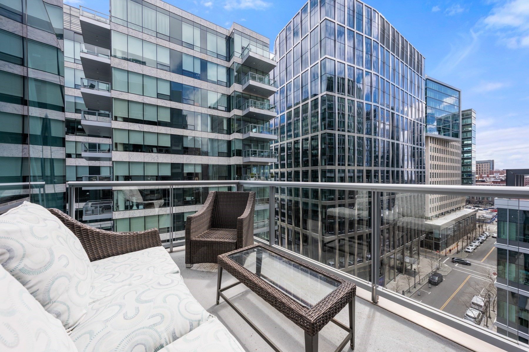 Furnished balcony with wicker seating overlooking modern glass buildings and city streets.