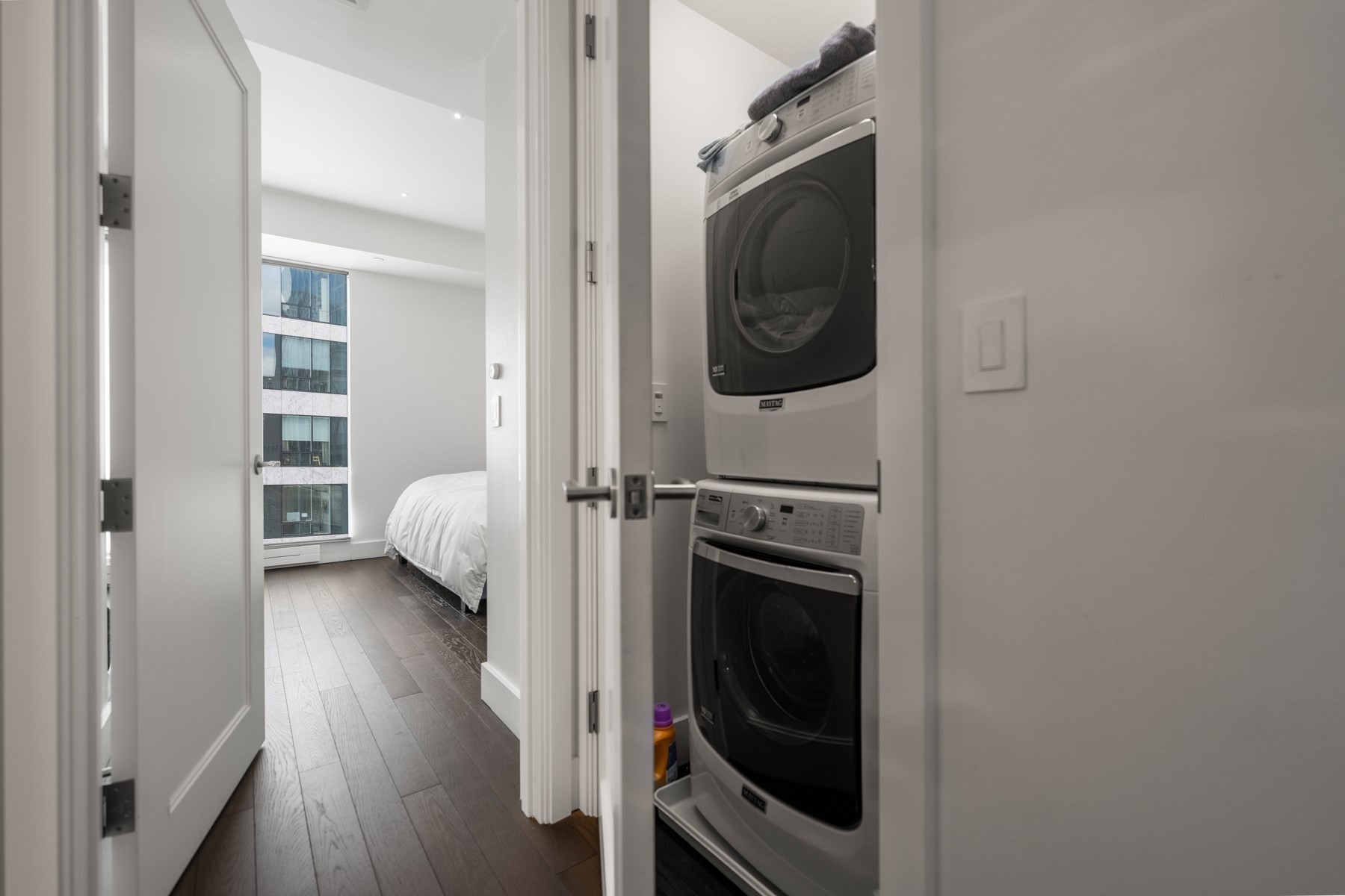 Stacked washer and dryer beside a bedroom with hardwood floors and a large window.