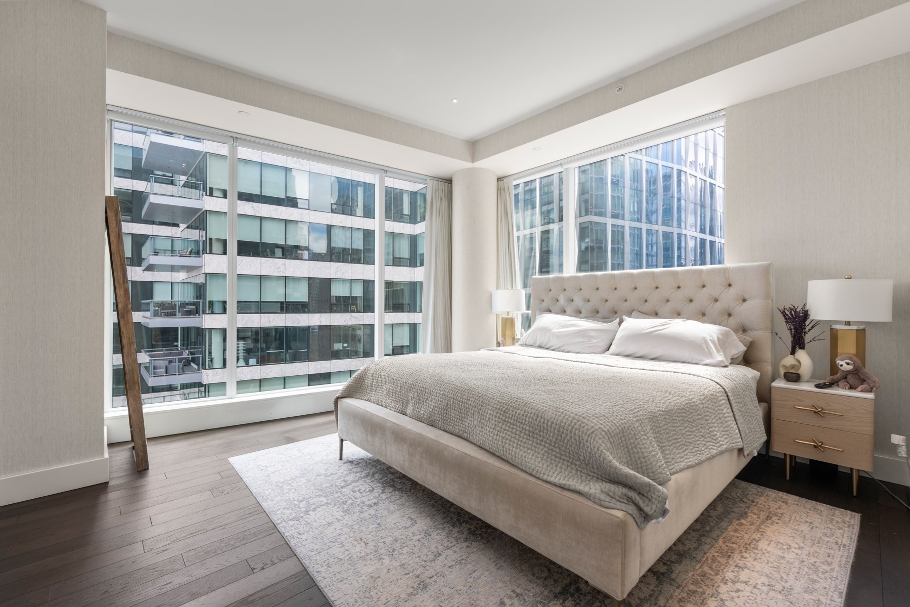 Furnished bedroom with a tufted bed and expansive corner windows overlooking modern city buildings.