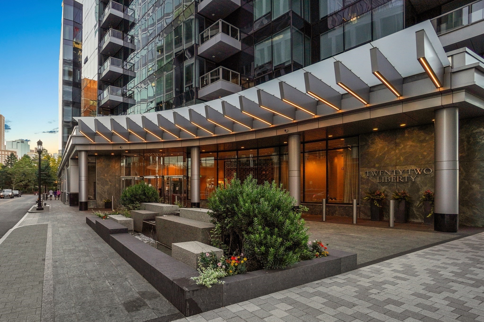 Twenty Two Liberty building entrance with glass balconies, an illuminated canopy, and landscaped planters.