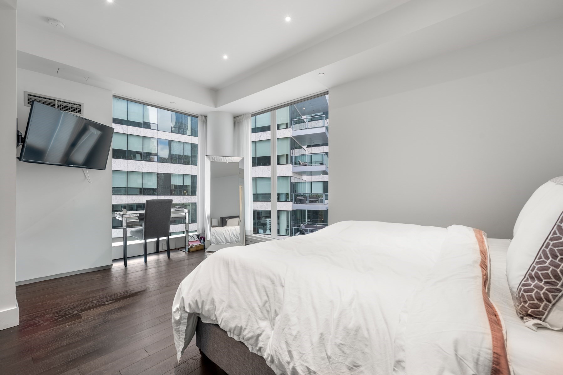 Modern bedroom with a white bed, dark wood floors, and corner windows overlooking neighboring buildings.