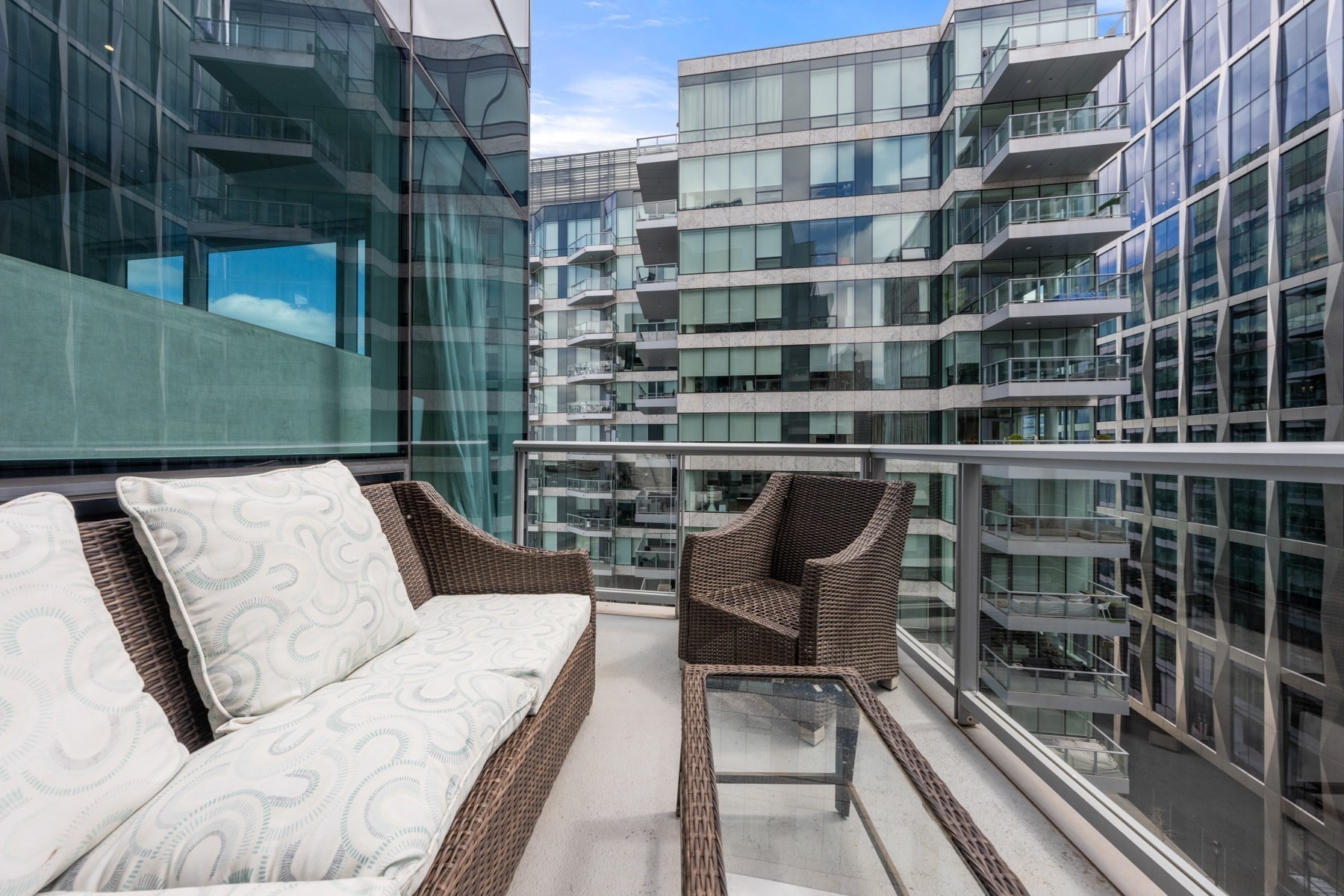 Apartment balcony with wicker seating and glass railing overlooking modern condominium buildings.