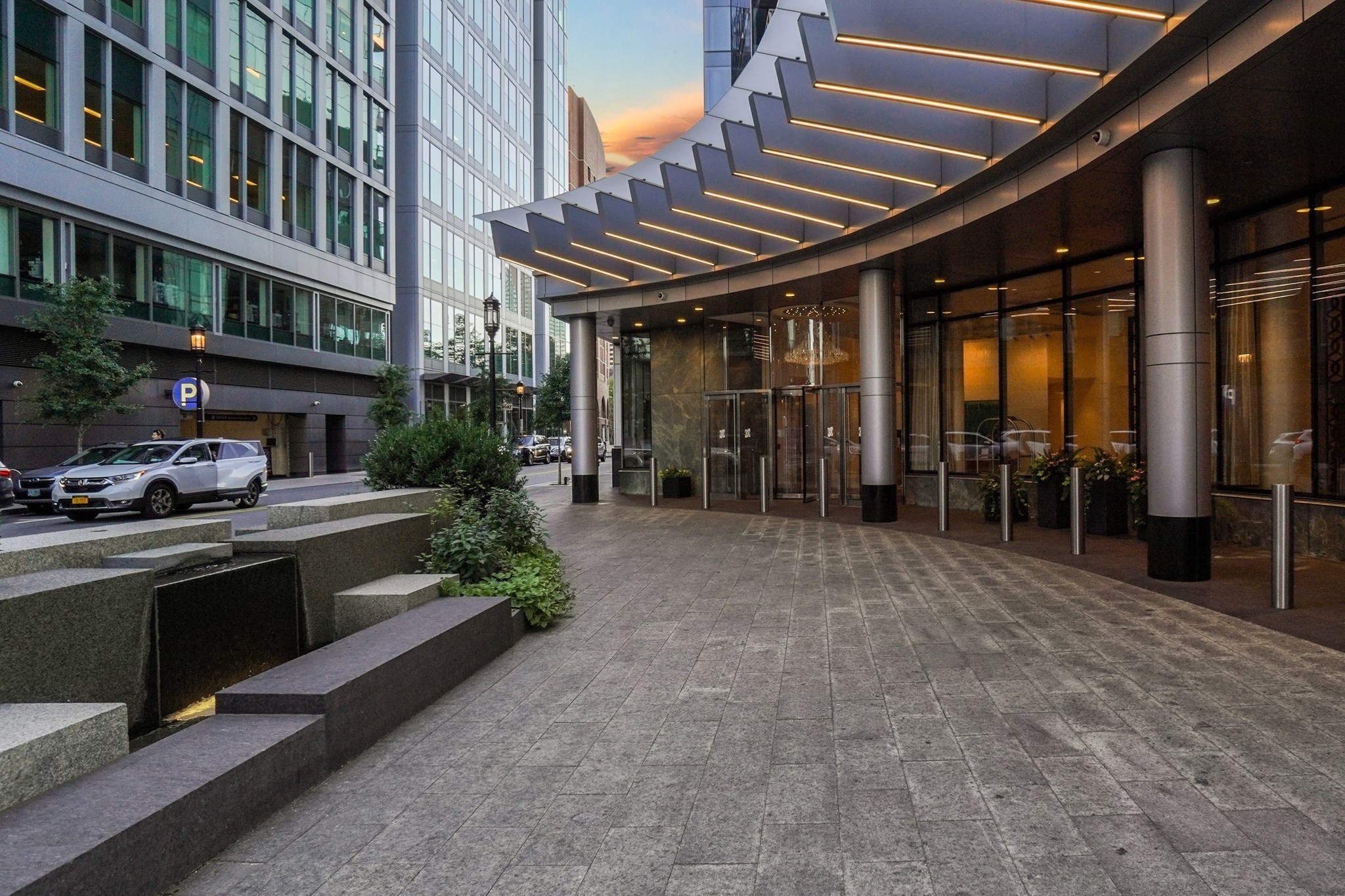 Modern high-rise entrance with a broad illuminated canopy and paved forecourt at dusk.