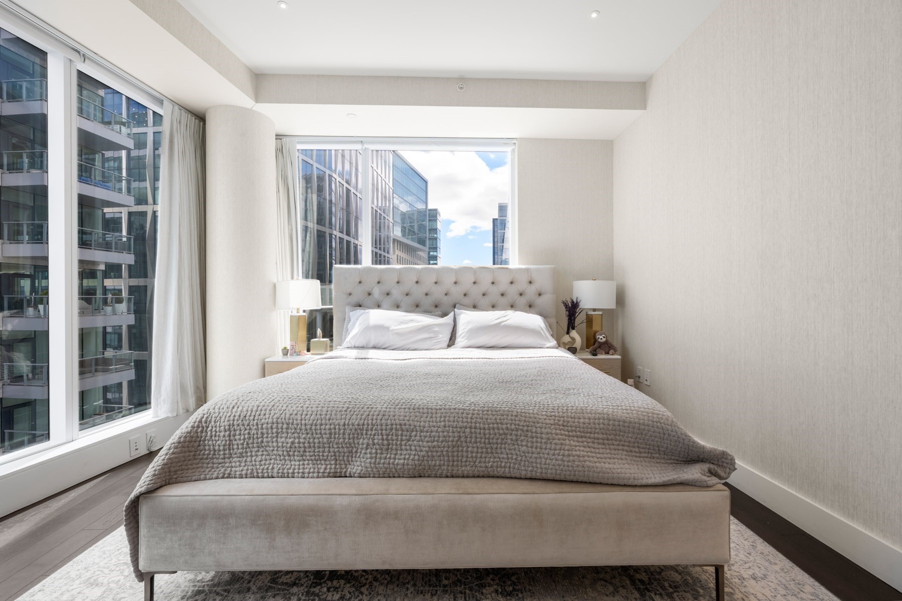 Bedroom with a tufted upholstered bed and floor-to-ceiling windows overlooking modern buildings.