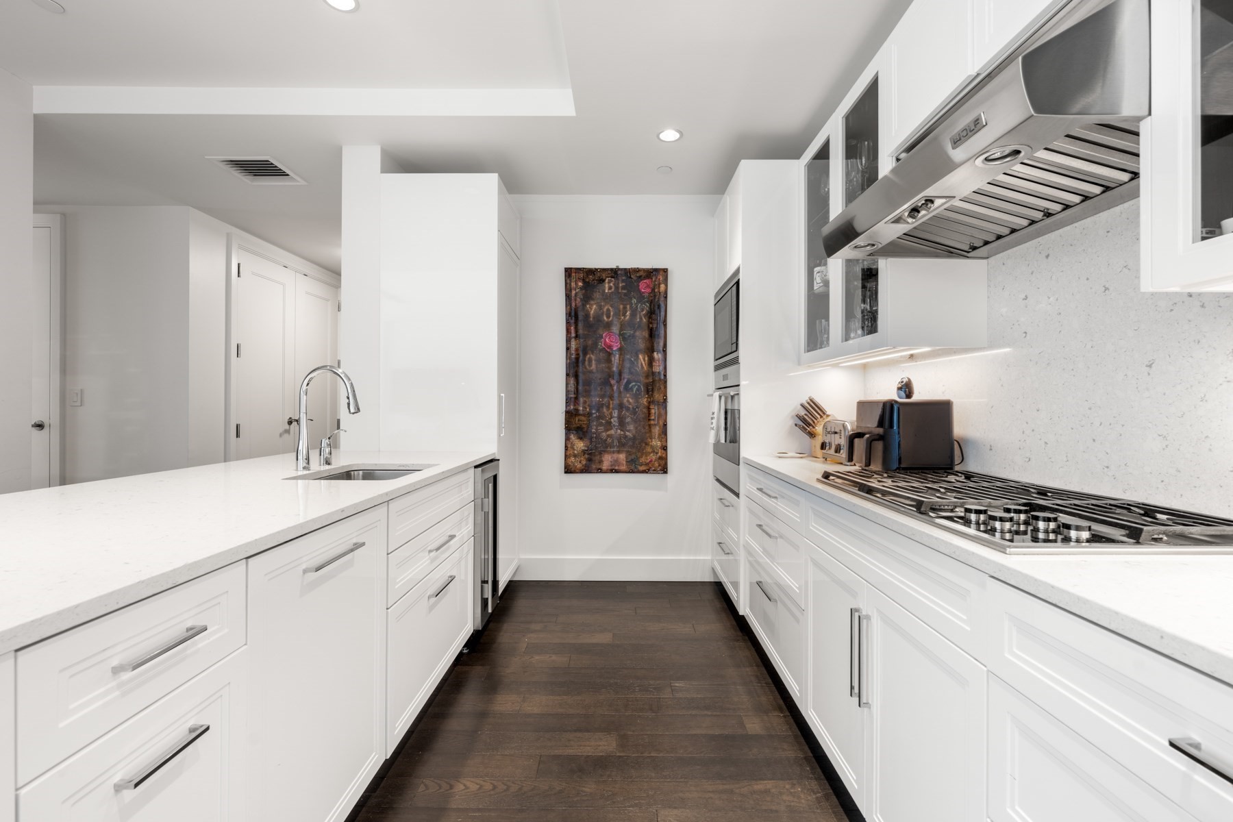 Modern white kitchen with a large island, stainless steel range hood, and dark wood flooring.