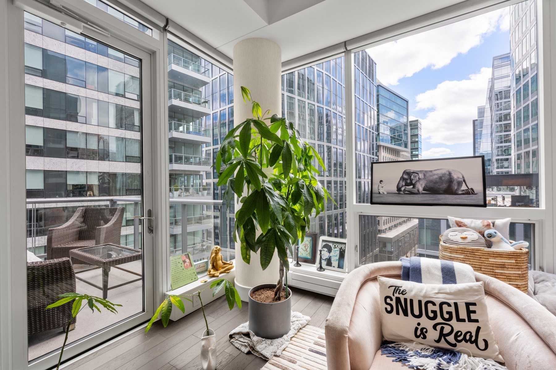 Furnished corner sitting area with floor-to-ceiling windows, a potted plant, and a balcony overlooking modern city buildings.