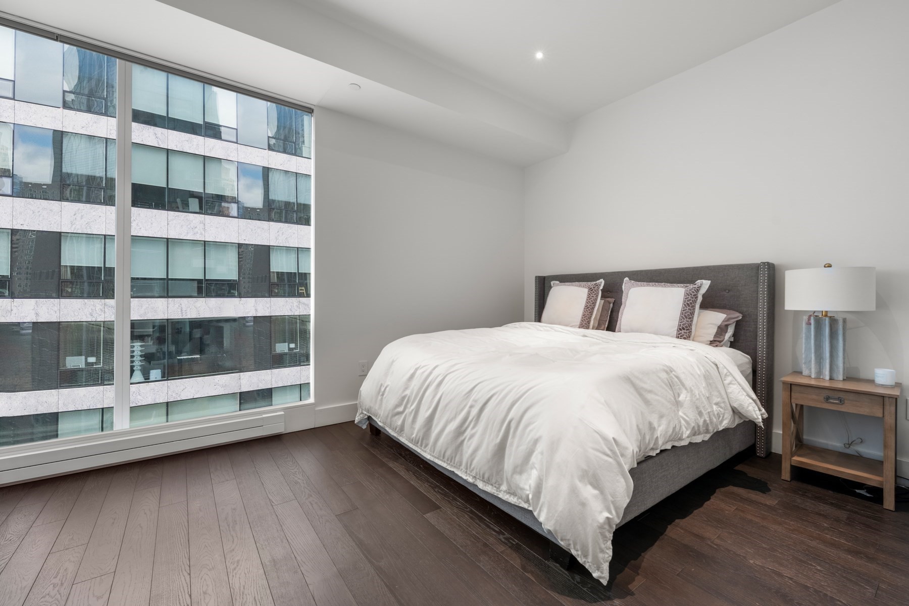 Bedroom with a large white bed, dark wood floors, and a floor-to-ceiling window overlooking a neighboring building.