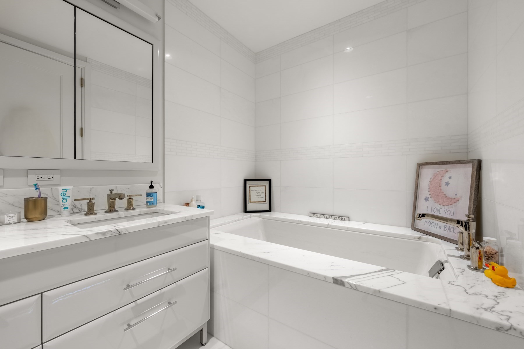White-tiled bathroom with a marble-edged bathtub, matching vanity, mirrored cabinet, and yellow rubber ducks.