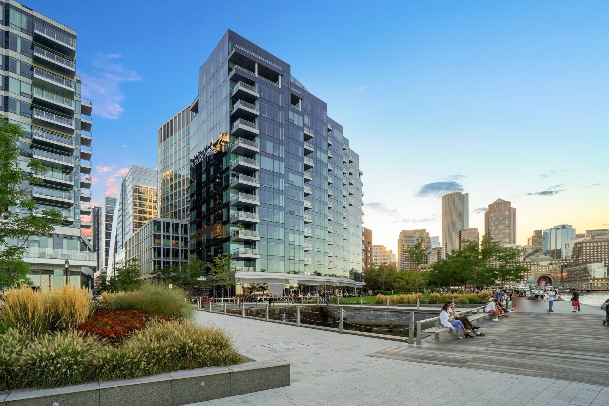Modern waterfront buildings beside a landscaped promenade and boardwalk, with the Boston skyline beyond.