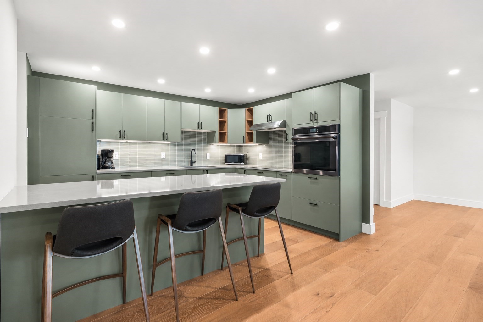 Modern kitchen with sage-green cabinets, a white peninsula, three bar stools, and light hardwood floors.
