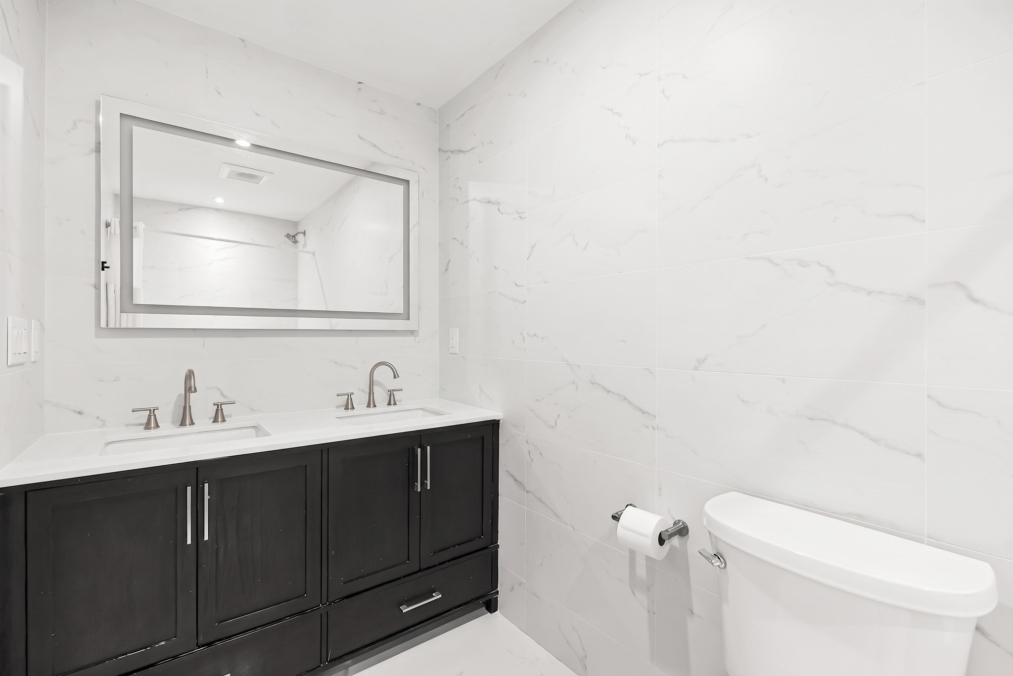 Bathroom with a dark double vanity, marble-look tile, wide mirror, and white toilet.